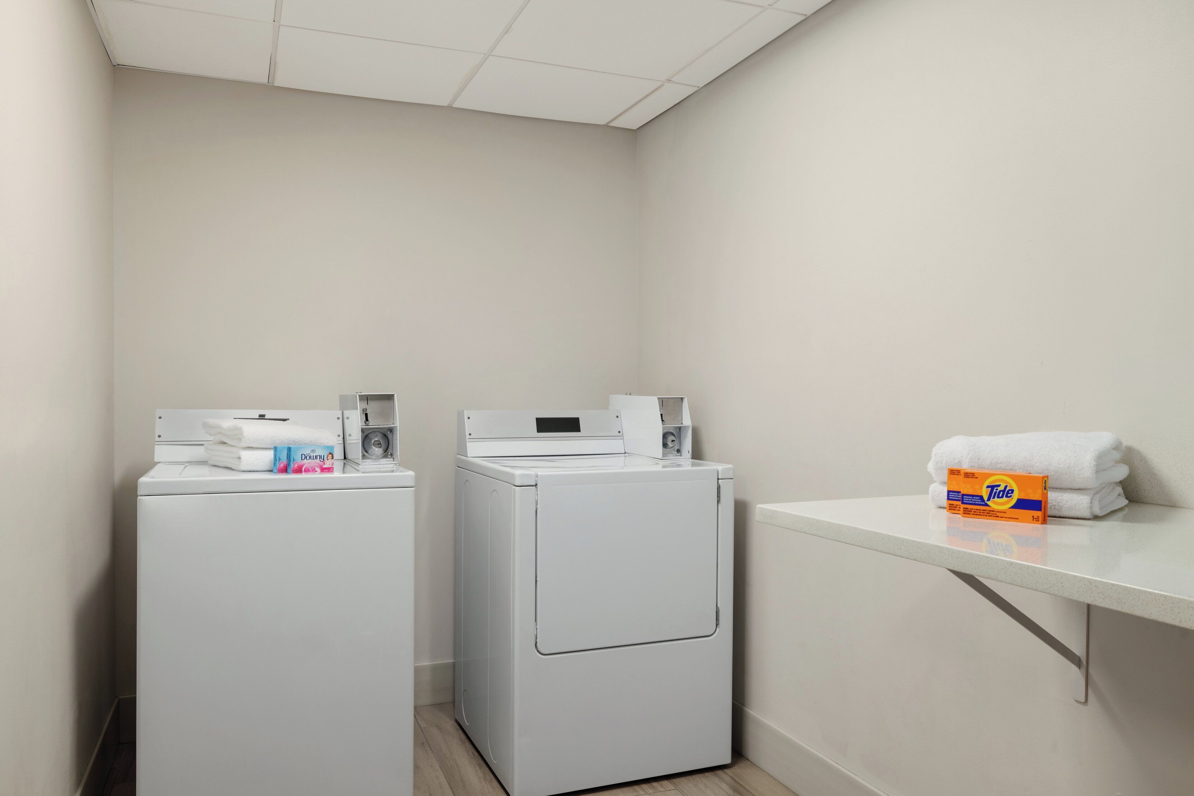 A laundry room with two white front-loading washing machines, a shelf with folded white towels, and a box of Tide detergent.