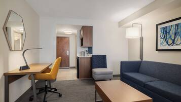 Et moderne hotelværelse med et skrivebord, en gul stol, en blå sofa, en lille skammel, en lampe og et billede på væggen.