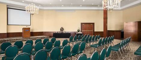 A conference room with rows of green chairs, a podium, a screen, and two chandeliers.