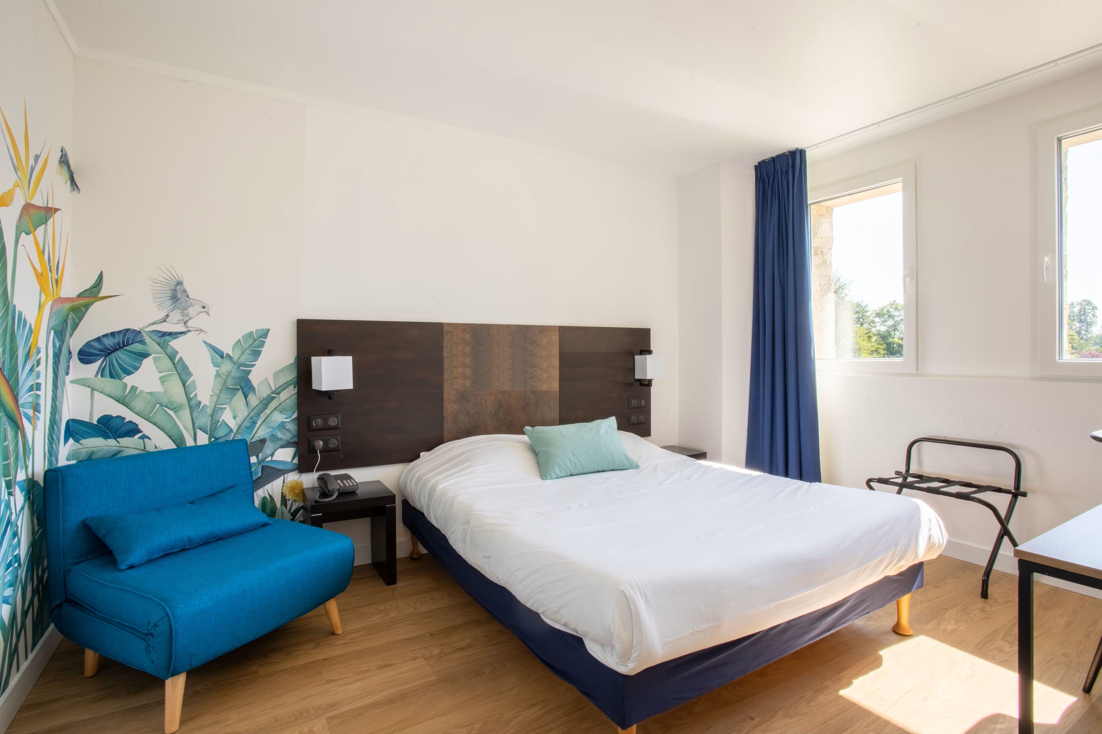 A hotel room with a bed, a blue chair, a nightstand, a desk, and a window with curtains.