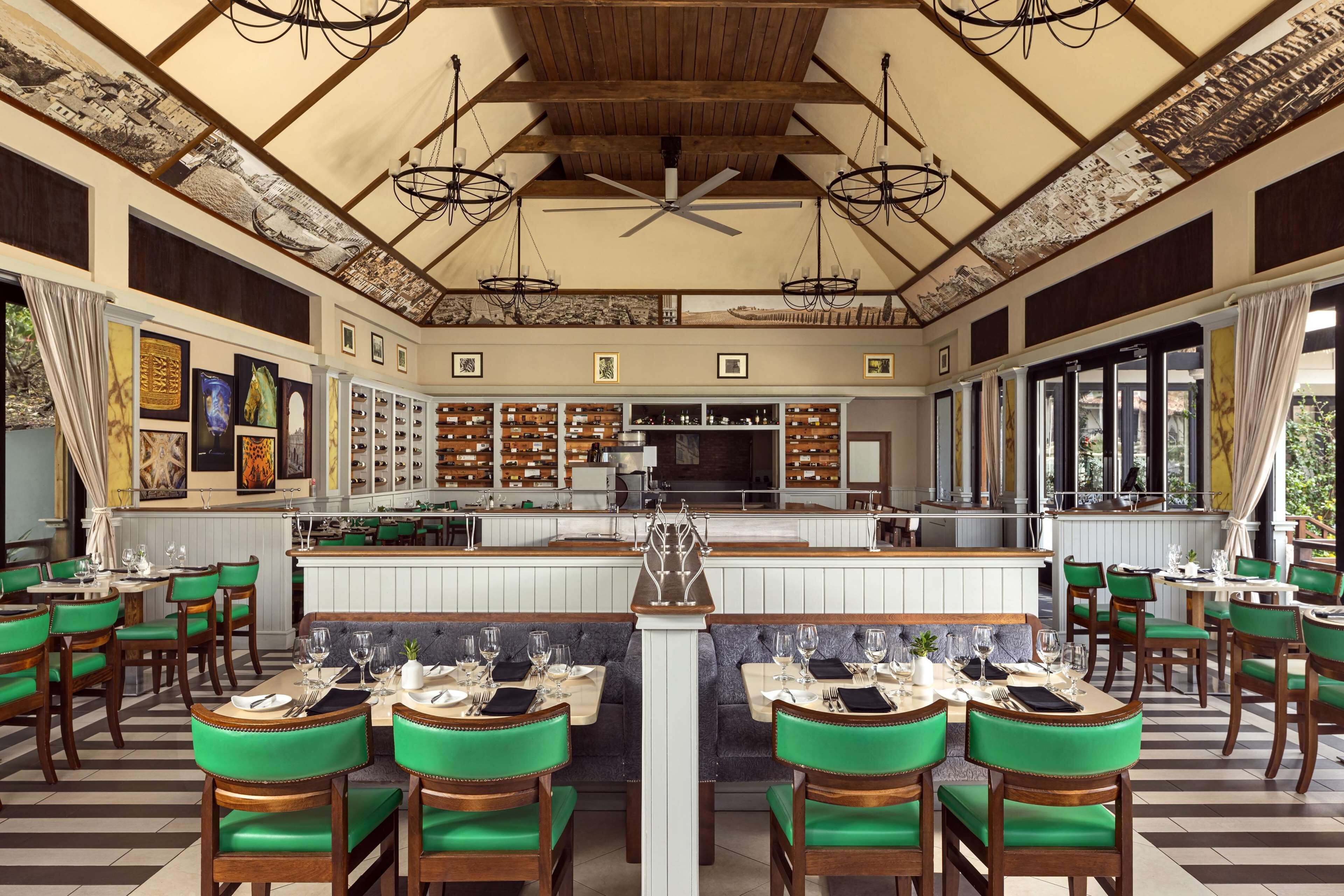 A restaurant with a central bar, green upholstered chairs, and a high ceiling with exposed beams.