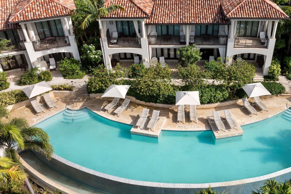 A large, curved swimming pool with multiple lounge areas and umbrellas, surrounded by lush greenery and a multi-story building with balconies.