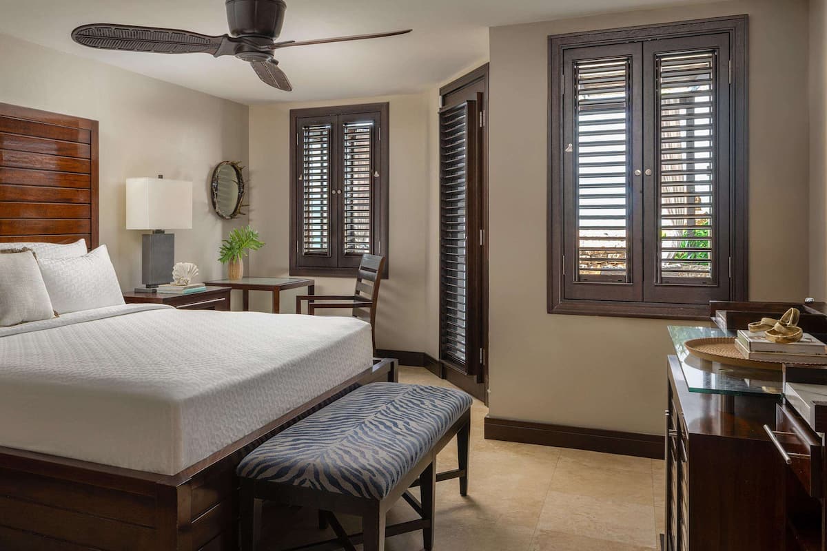 A bedroom with a large bed, a desk, and a chair. There is a ceiling fan, a lamp, and a window with shutters.