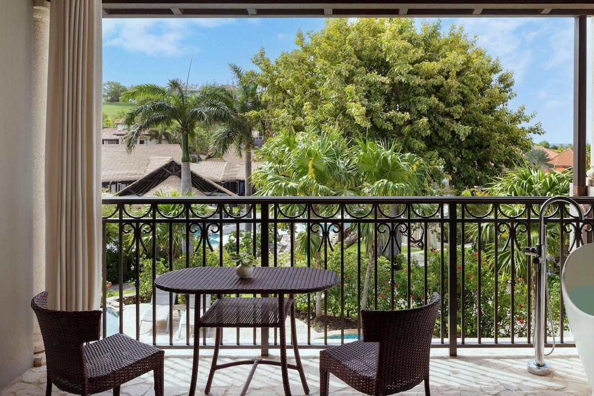 A balcony with wicker furniture, a white bathtub, and a view of a lush garden and buildings.
