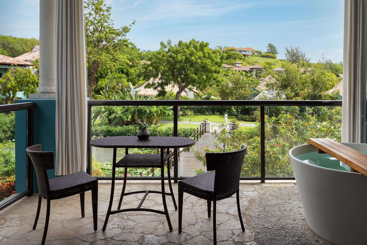 A balcony with a view of a lush garden and houses, a round table with chairs, and a bathtub.