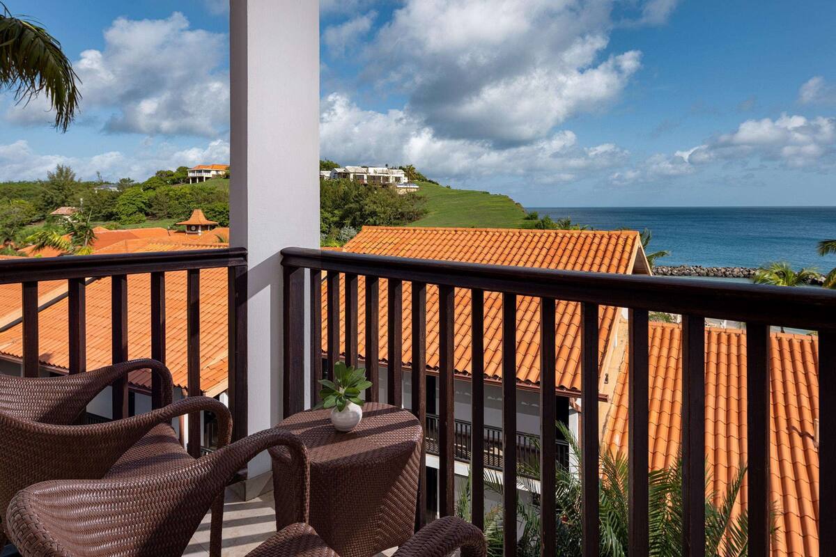 A balcony with wicker chairs and a potted plant, overlooking a coastal area with terracotta-roofed buildings and a view of the ocean.