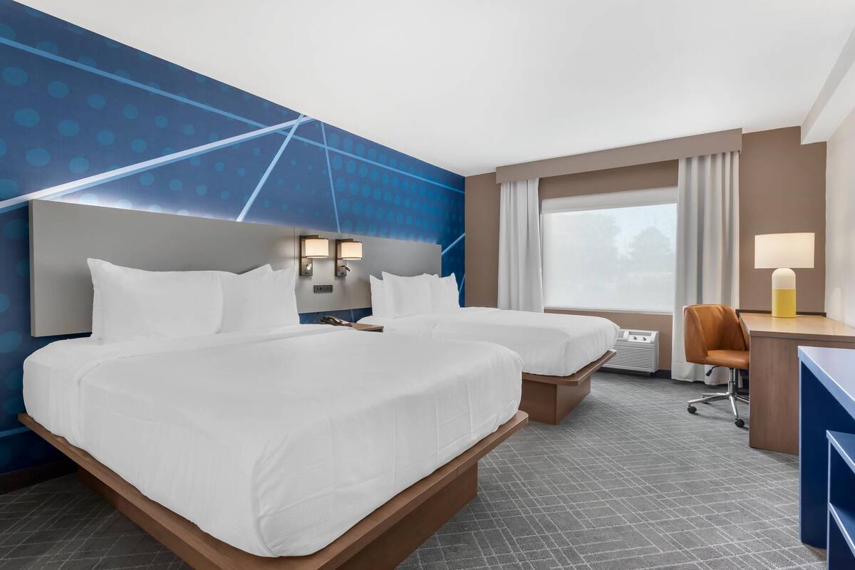Quarto de hotel com duas camas, uma escrivaninha, uma cadeira, uma janela com cortinas e uma parede com padrão geométrico.