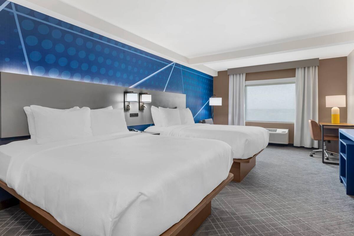 Quarto de hotel com duas camas, uma mesa, uma cadeira, um abajur e uma janela grande.