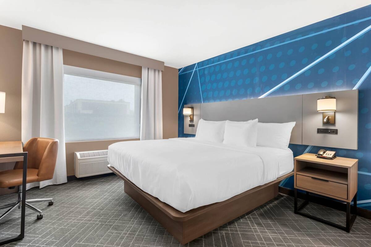 Quarto de hotel com uma cama grande, uma escrivaninha, uma cadeira, um criado-mudo e uma luminária fixada na parede.