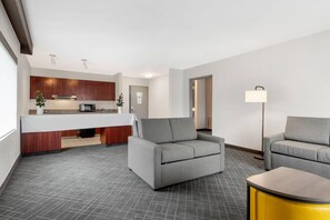 Una habitación de hotel moderna con kitchenette, dos sofás grises, una mesita auxiliar amarilla y una lámpara de pie.
