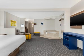 Habitación de hotel con cama, escritorio, silla, pequeño banco, bañera, televisor y espejo adosado a la pared.