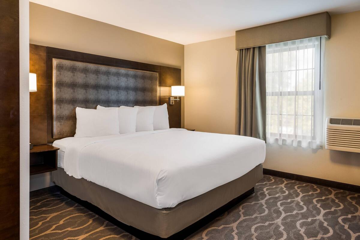 A hotel room with a large bed, a headboard, two wall-mounted lamps, a window with curtains, and a patterned carpet.