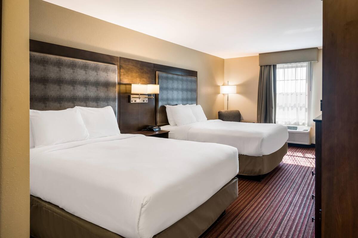 A hotel room with two beds, a large headboard, bedside lamps, a nightstand, and a view of the outdoors through a window.