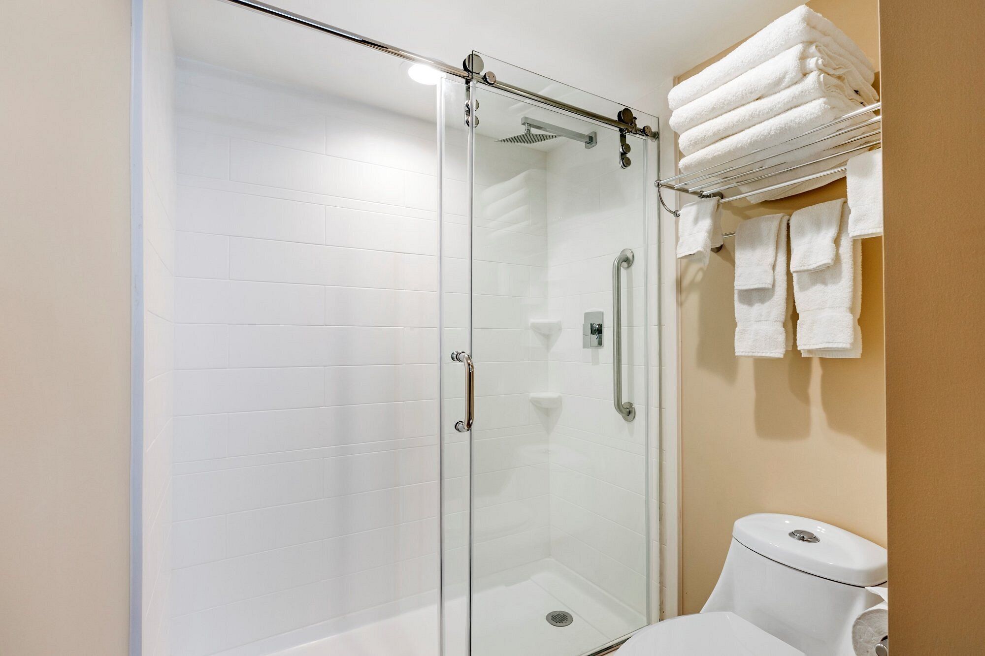 A glass-enclosed shower with a white tiled wall, a toilet with a beige wall, and a towel rack with folded towels.