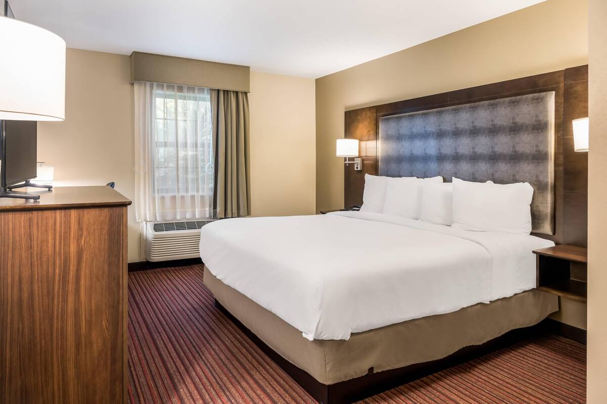 A hotel room with a large bed, a desk, a lamp, a window with curtains, and a wall-mounted light.