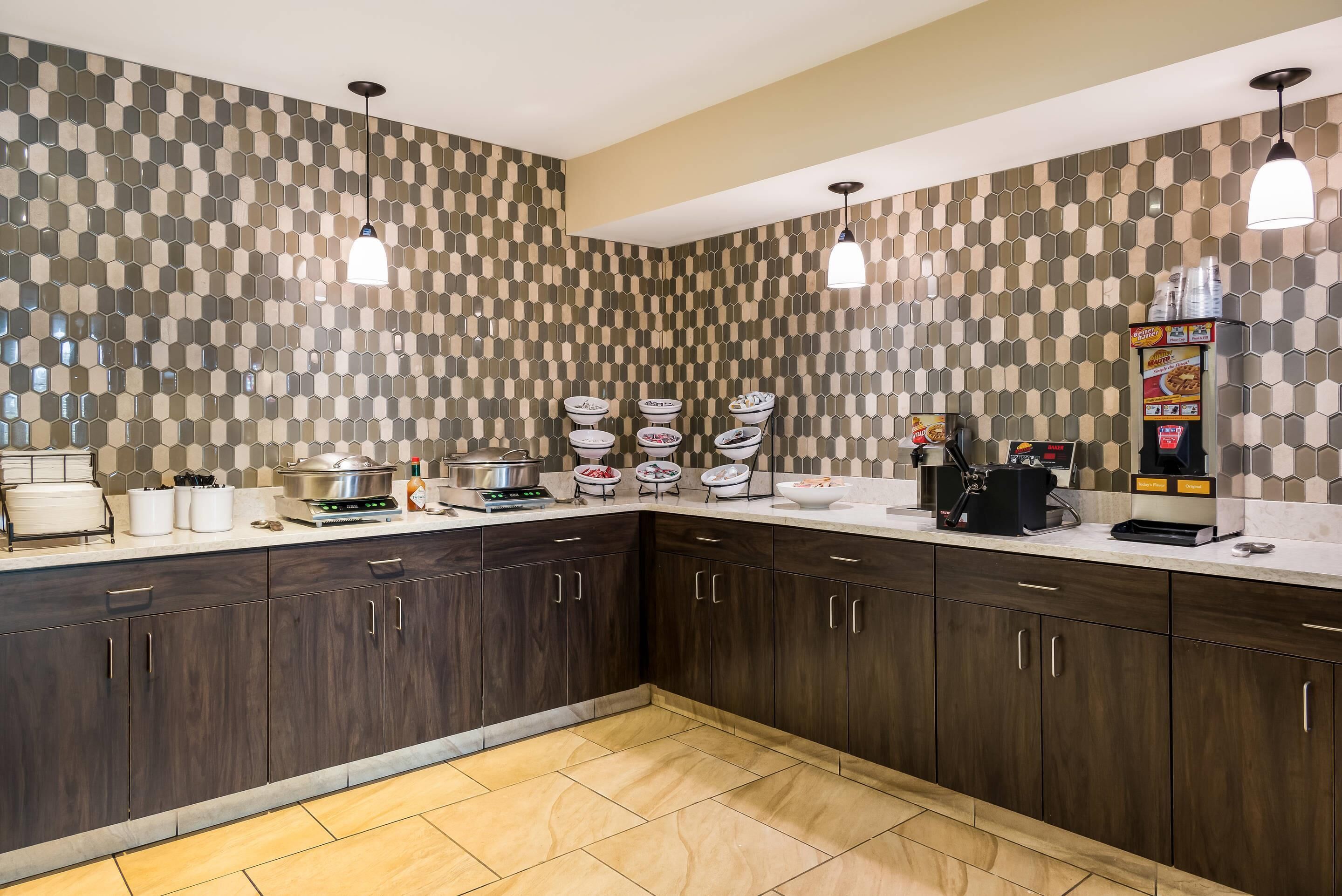 A modern kitchen with dark wood cabinets, a white countertop, and a patterned backsplash. There are two pendant lights, a coffee maker, and a box of cereal on the counter.