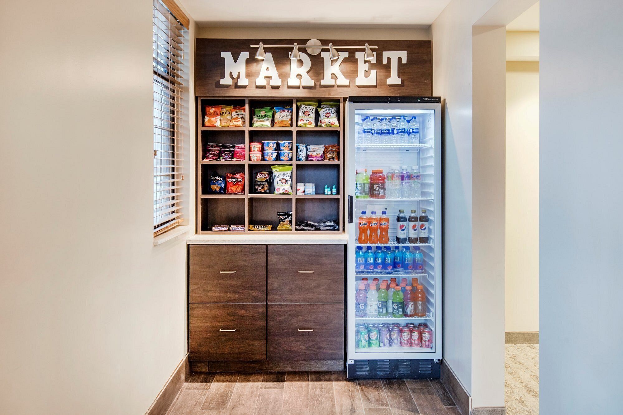 A kitchenette with a built-in mini-fridge stocked with beverages and snacks, a shelf with packaged food items, and a wooden cabinet below.