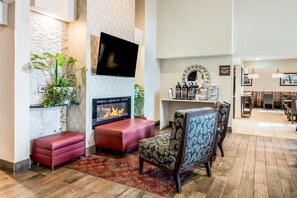 A modern living room with a fireplace, red leather ottoman, patterned armchair, and a flat-screen TV mounted on the wall.