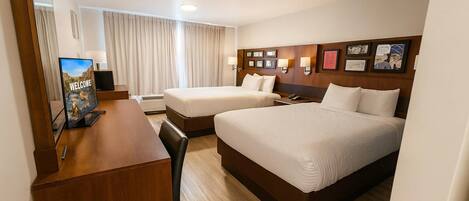 A hotel room with two beds, a desk, a TV, and a large window with curtains.
