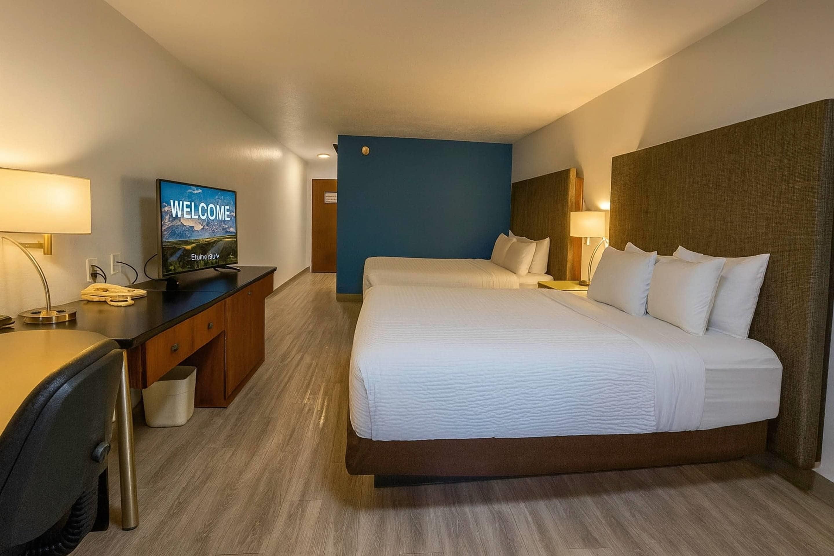 Habitación de hotel con una cama grande, un escritorio con una computadora y un televisor que muestra la palabra “WELCOME” en la pantalla.