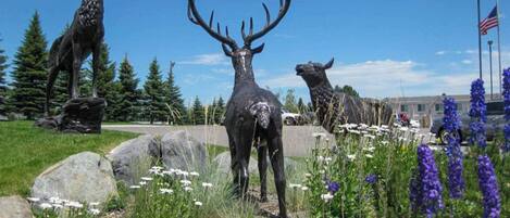 A statue of a deer in a park with trees and flowers.