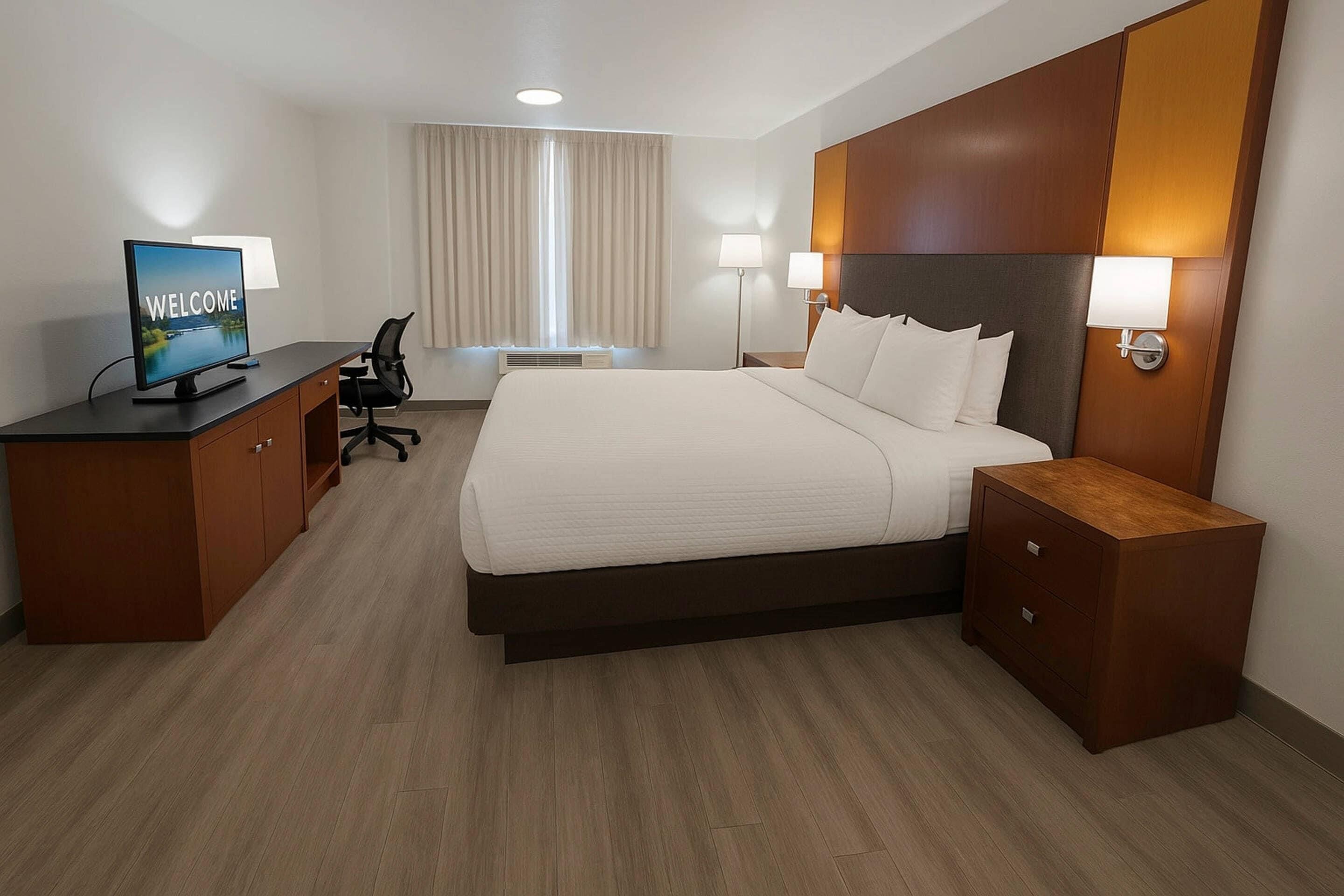 A hotel room with a large bed, a desk with a computer, a chair, two bedside lamps, and a window with curtains.