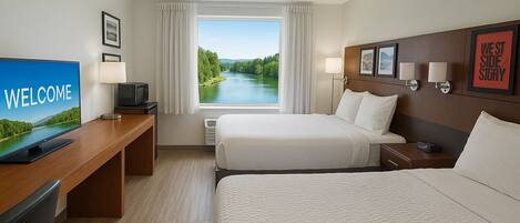 Una habitación de hotel con una cama, un escritorio con un televisor mostrando “BIENVENIDOS”, una ventana con vistas a un río y una pared con fotografías enmarcadas y un letrero que dice “WEST SIDE”.
