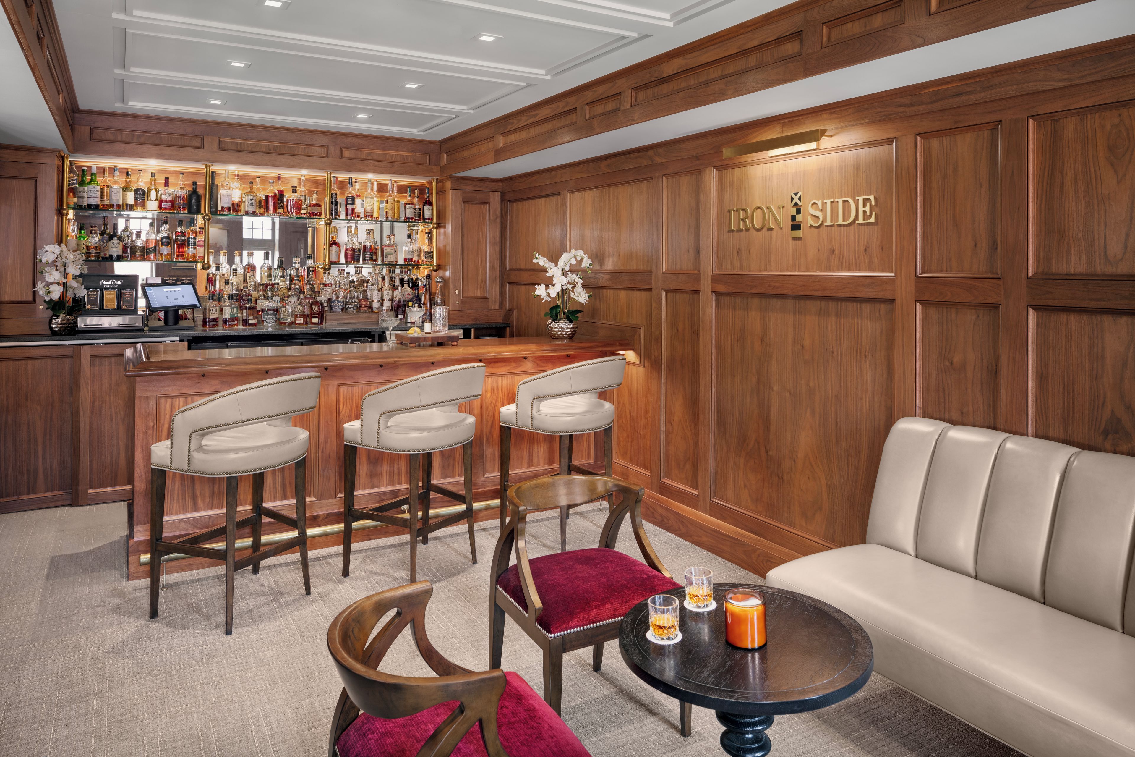 A bar area with a wooden bar counter, multiple liquor bottles, and a sign reading 'TRON SIDE'. There are three bar stools and a sofa in the seating area.