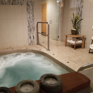 A hot tub area with a tiled floor, a wooden bench, a potted plant, and towels on a rack.