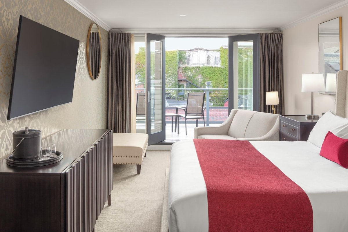 Habitación de hotel con cama, escritorio, televisor, balcón con silla y una mesa de comedor.