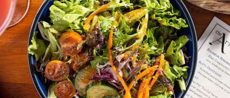 Une table avec un saladier bleu rempli de salade, une assiette avec de la viande grillée et du fromage, et un menu.