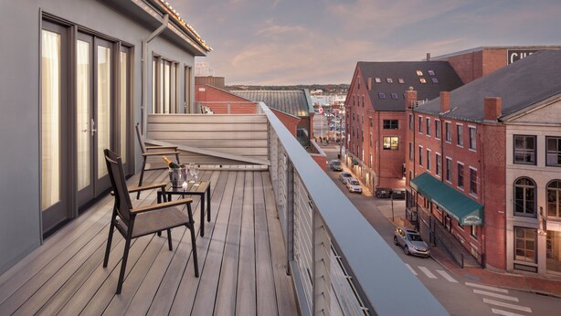 Een balkon met een tafel en stoelen, met uitzicht op een straat met gebouwen en geparkeerde auto's.