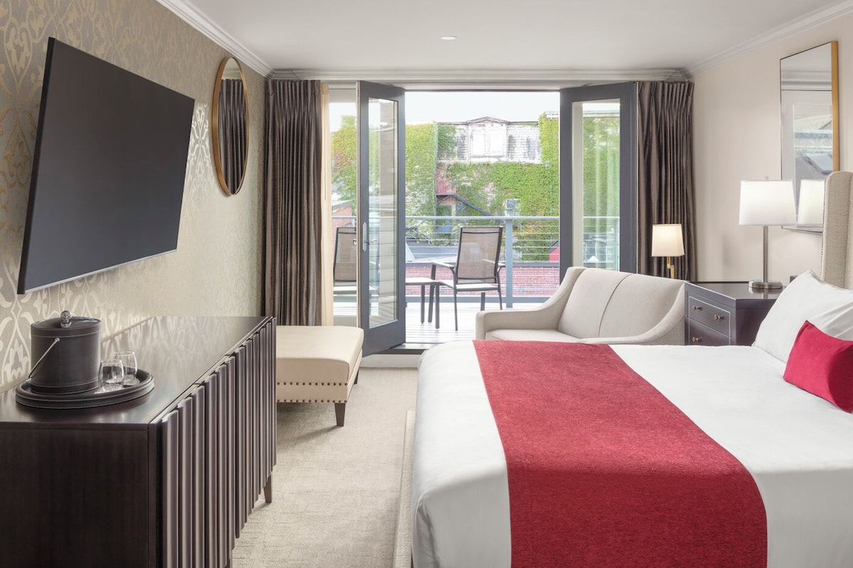 A hotel room with a bed, a desk, a TV, a balcony with a chair, and a dining table.