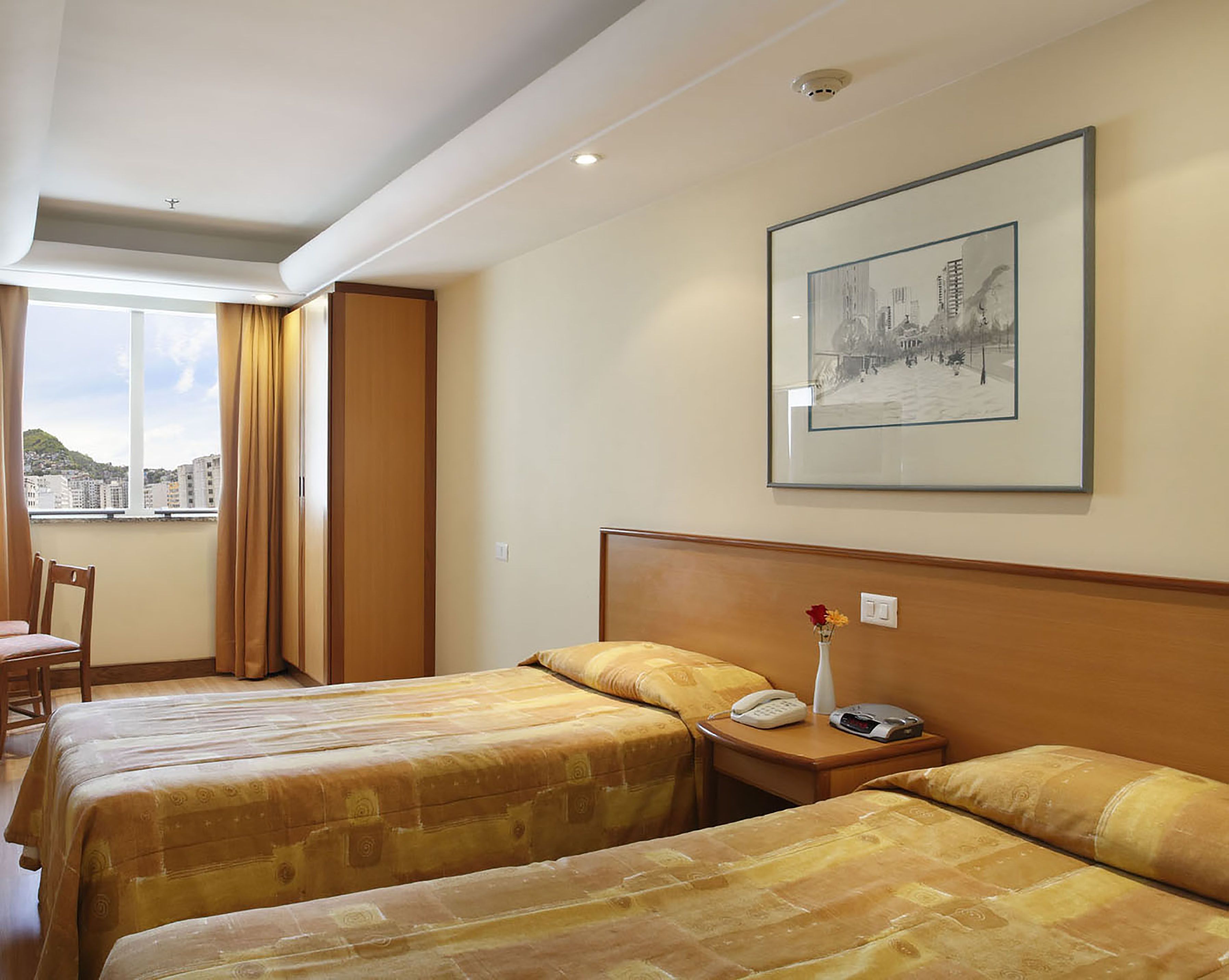 Une chambre d’hôtel avec deux lits, une vue sur la ville, un tableau encadré au mur et une table de chevet avec un téléphone et un vase de fleurs.
