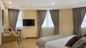 Habitación de hotel con cama, escritorio con desayuno, televisor y ventanas grandes con cortinas.