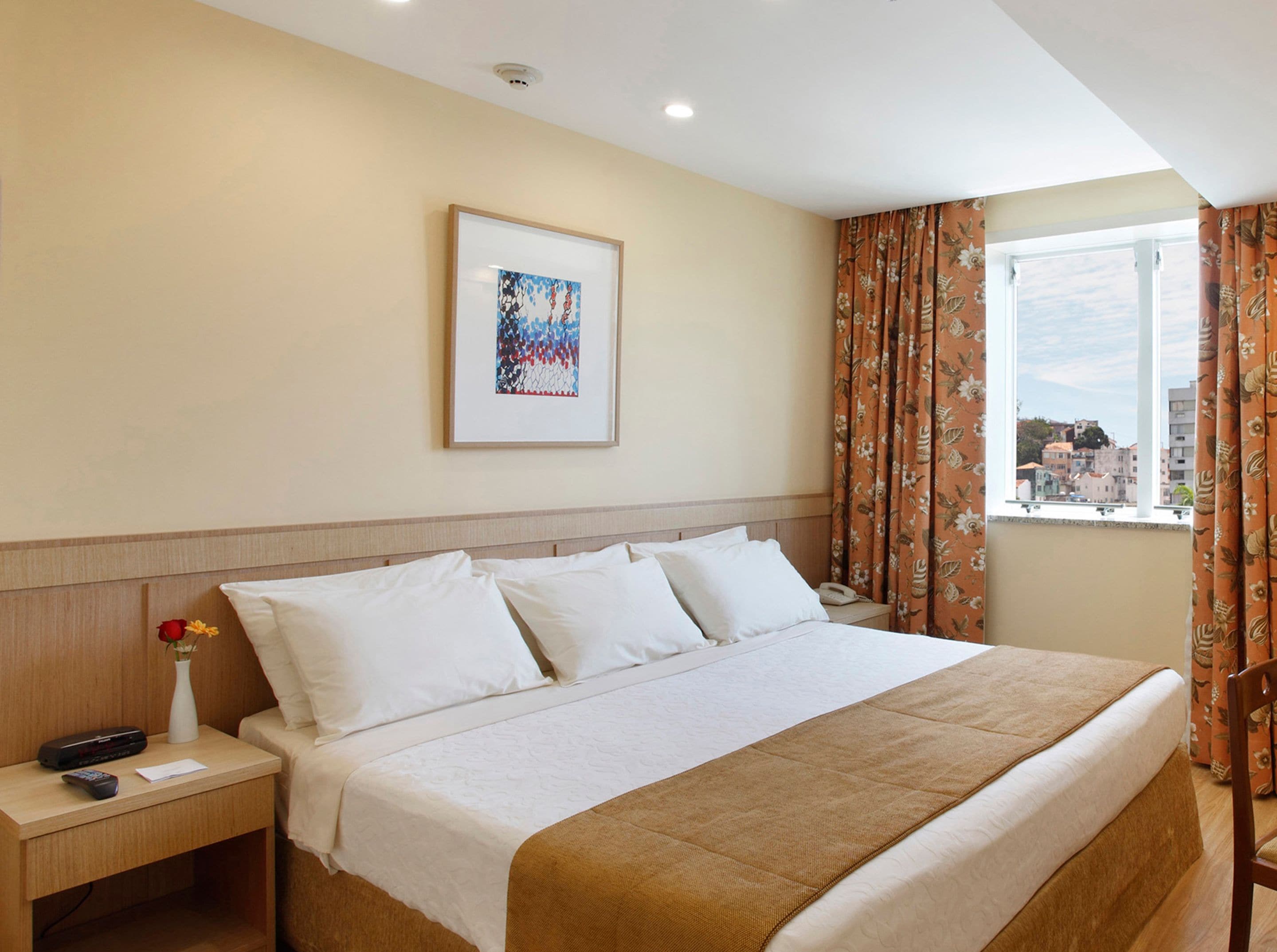 A hotel room with a large bed, a bedside table with a phone and remote control, a framed picture on the wall, and a window with curtains.