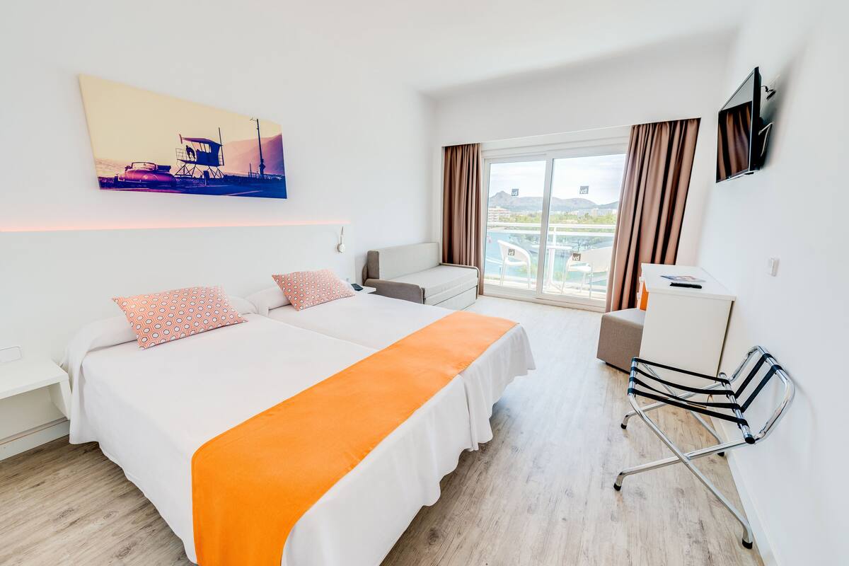 Una habitación de hotel con cama, escritorio, silla, televisión y ventana con cortinas.