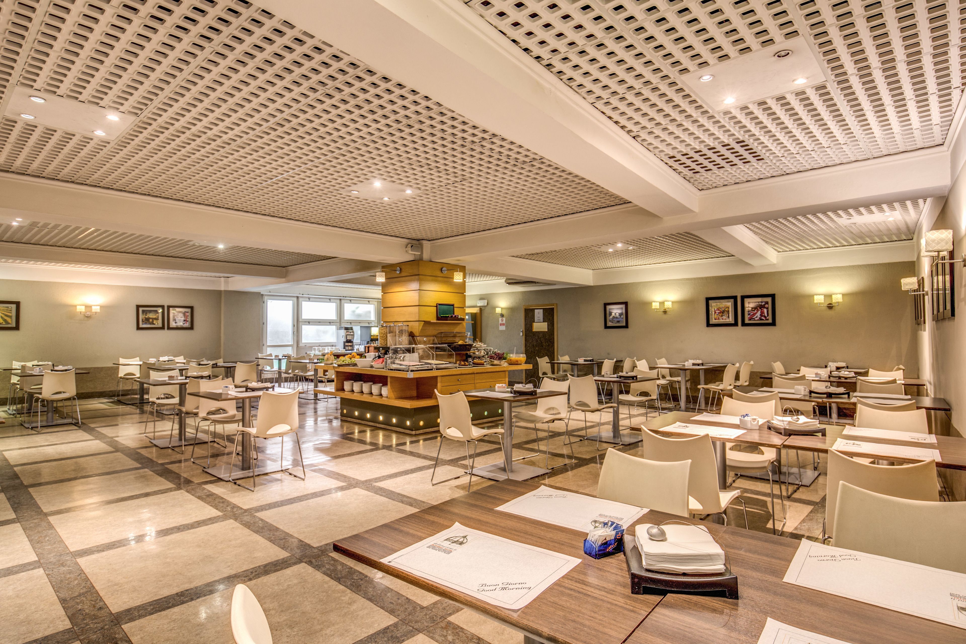 A spacious hotel dining area with multiple tables and chairs, a central bar, and a patterned ceiling.