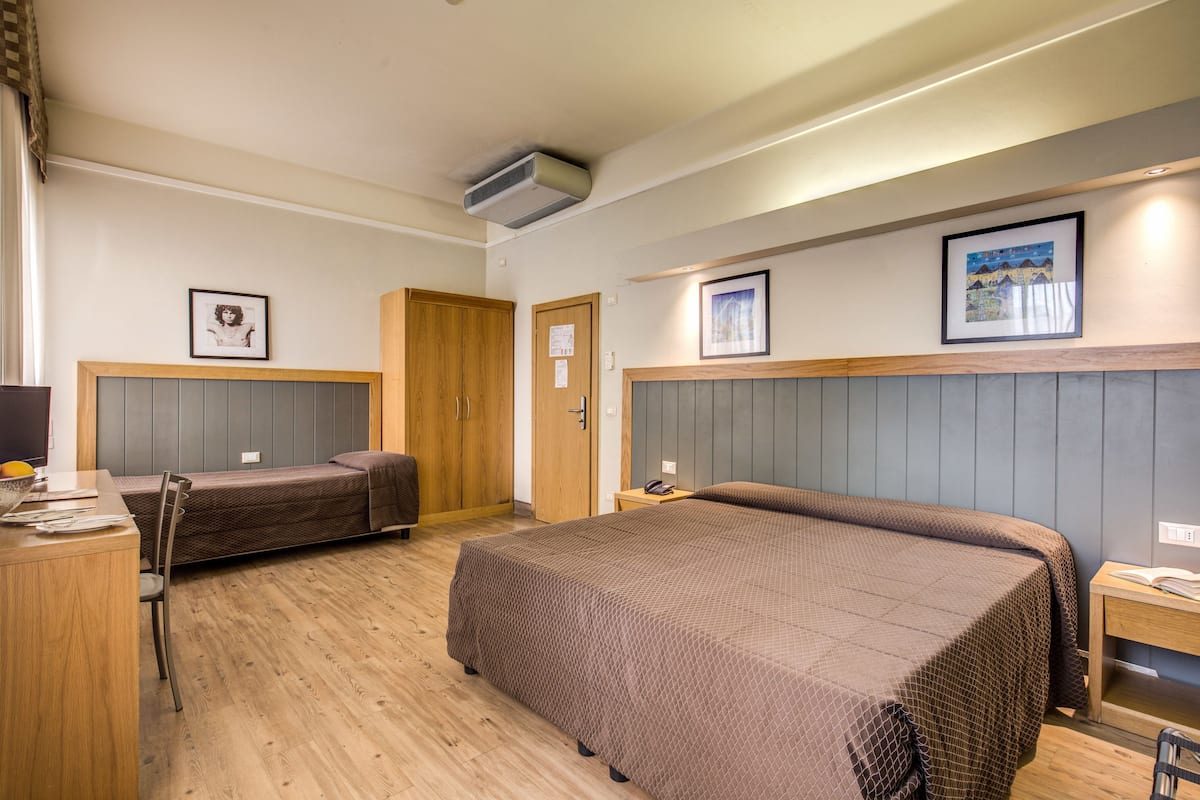 Hotellihuoneessa on suuri sänky, pienempi sänky, työpöytä ja tuoli. Seinällä on kehystettyjä tauluja ja katossa ilmastointilaite.