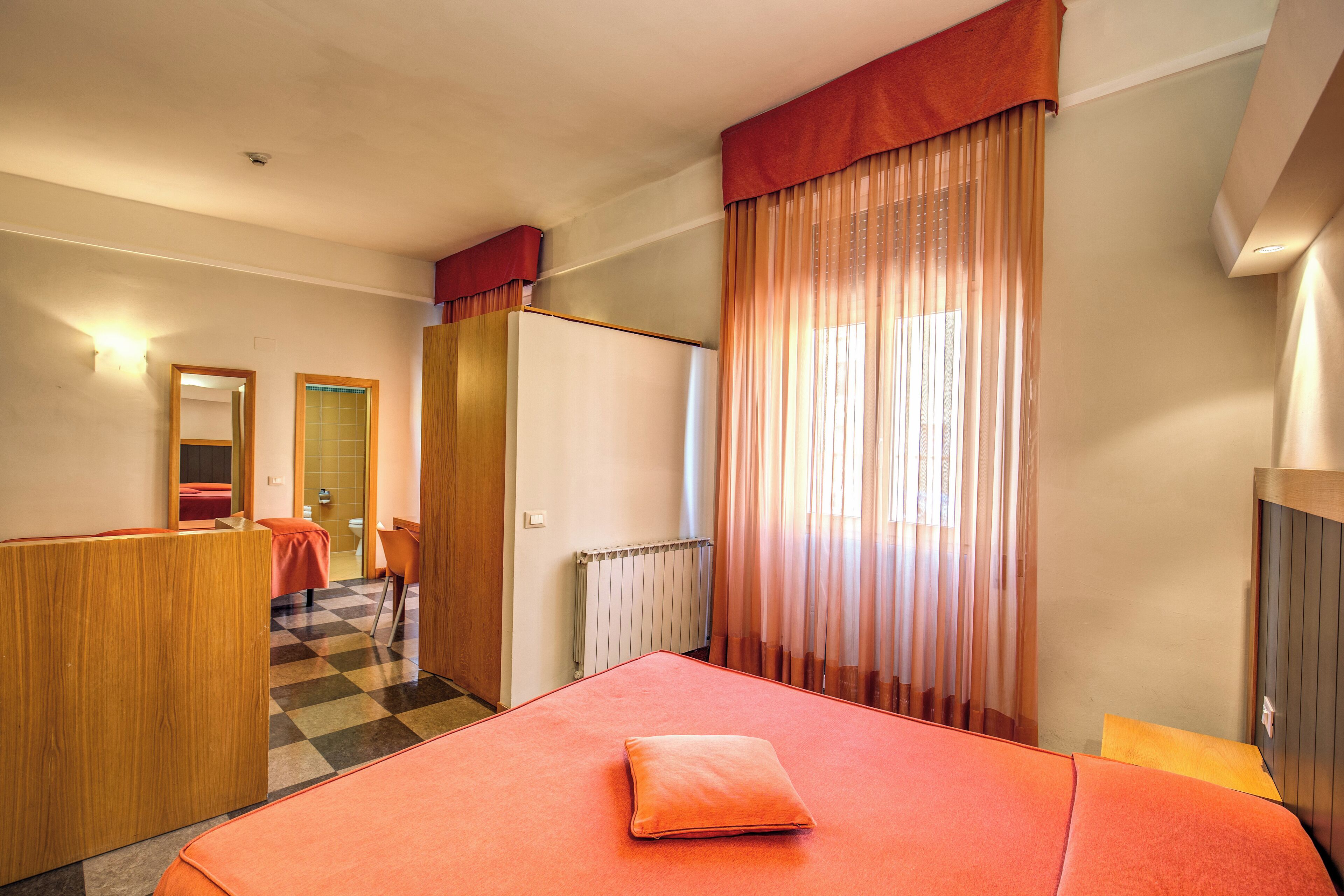 A hotel room with a checkered floor, a bed with an orange bedspread and pillow, a wooden desk, a mirror, and a window with sheer curtains.