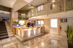 A hotel reception area with a marble floor, a reception desk, a staircase, and a second-floor balcony.