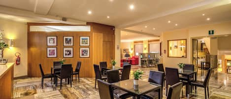 Un vestíbulo de hotel con suelo de mármol, varias mesas de comedor con sillas, un mostrador de recepción y cuadros en la pared.