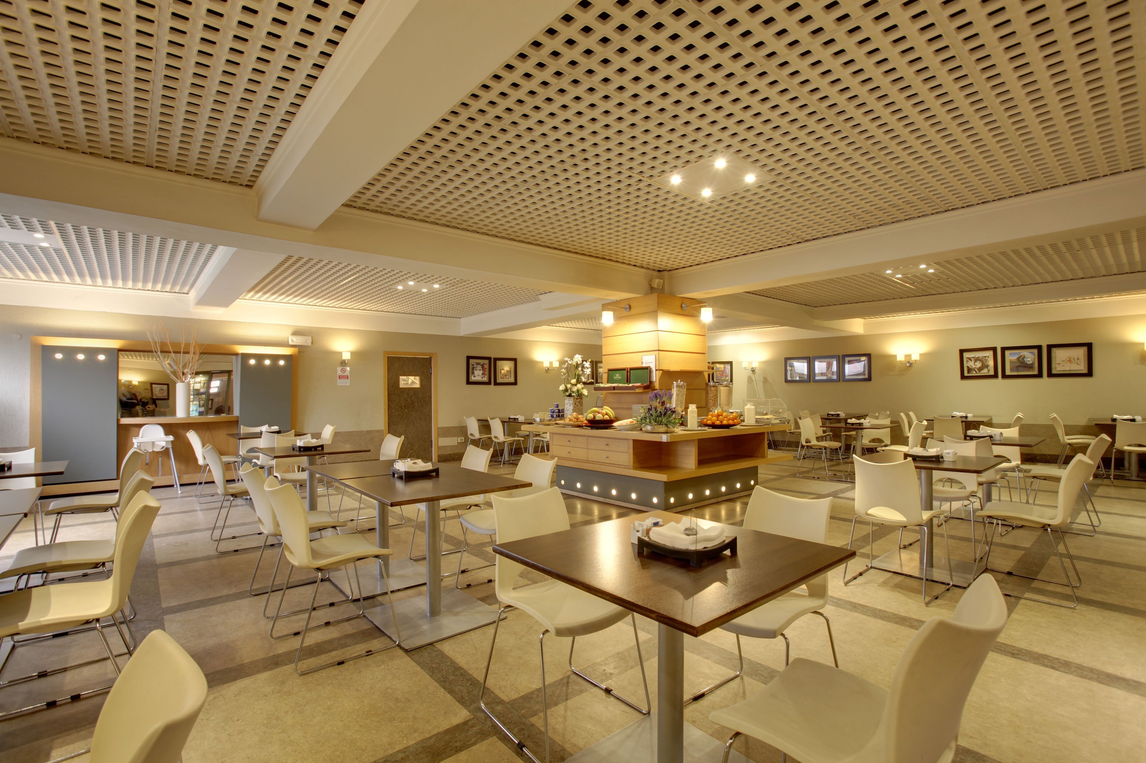 A hotel dining area with multiple tables and chairs, a central island with a fruit bowl and flowers, and a ceiling with a grid pattern.