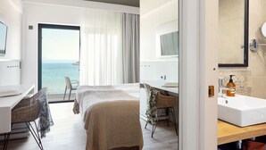 Habitación de hotel con cama, escritorio, silla, baño con lavamanos y espejo, y vista al mar a través de un ventanal grande.