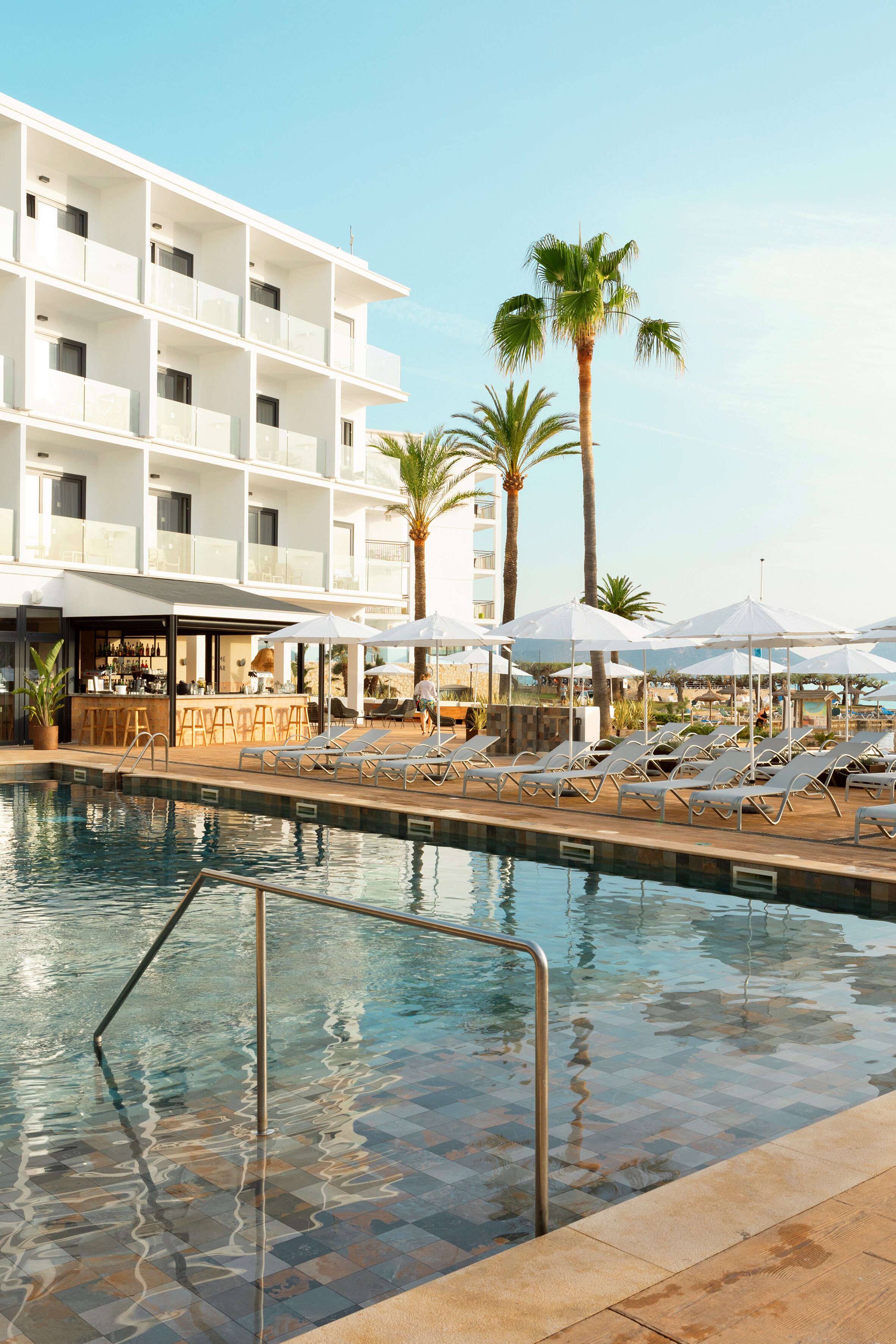 Zona de piscina de hotel con sillones de descanso, sombrillas y un bar.