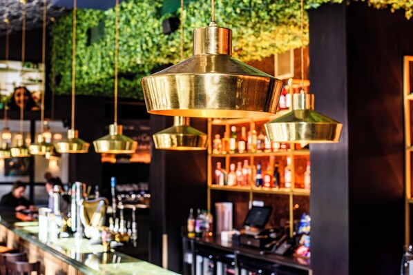 Zona de bar con lámparas colgantes de latón, estantes con botellas y una pared cubierta de follaje verde.