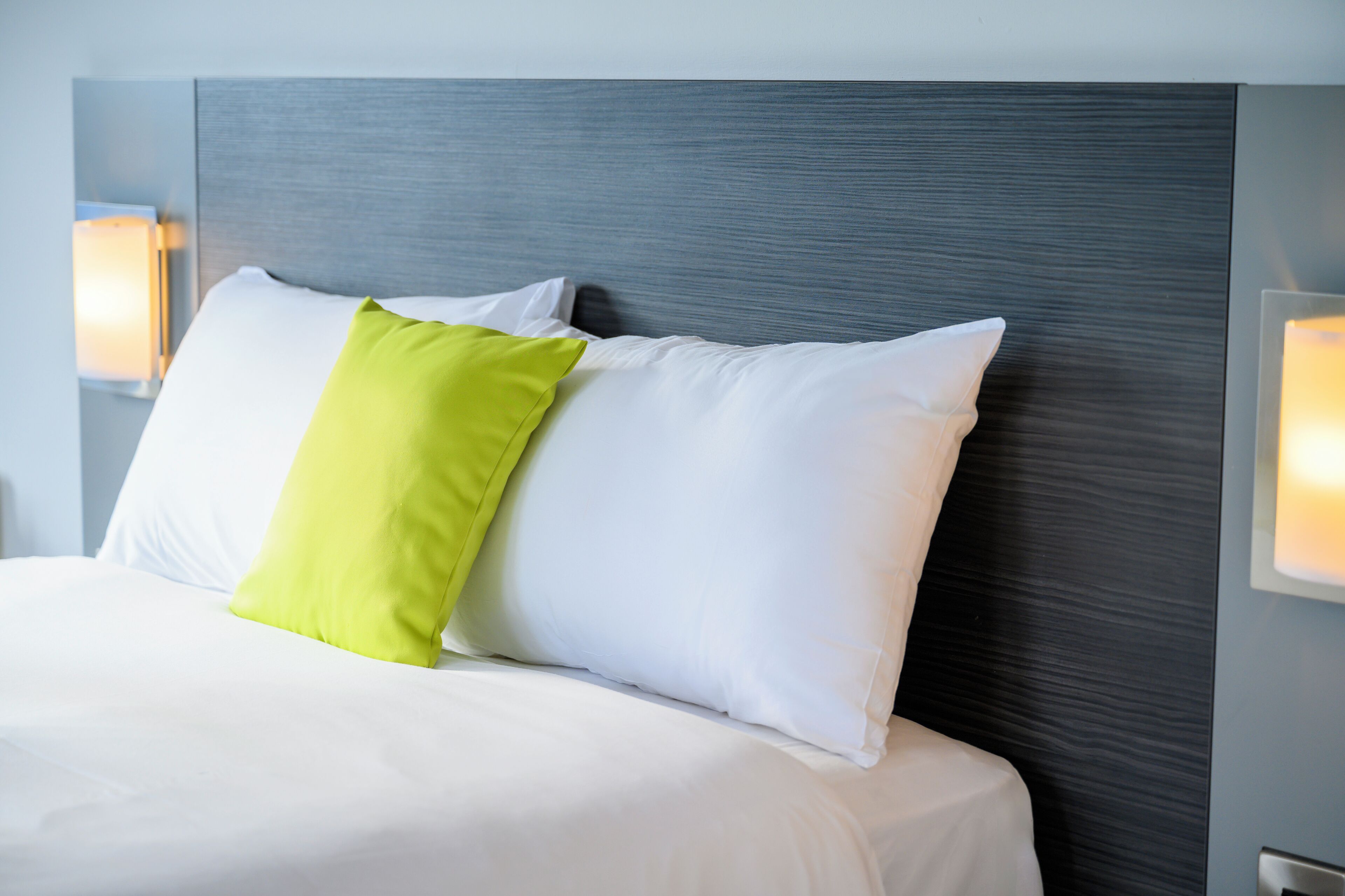 A neatly made bed with white and green pillows, a grey headboard, and wall-mounted reading lamps.