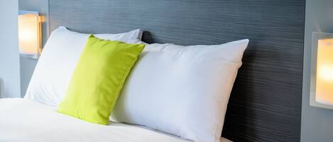 A neatly made bed with white and green pillows, a grey headboard, and wall-mounted reading lamps.