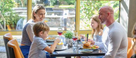 Una familia cenando en una mesa con ventanas amplias, vistas a la vegetación y una fuente al fondo.