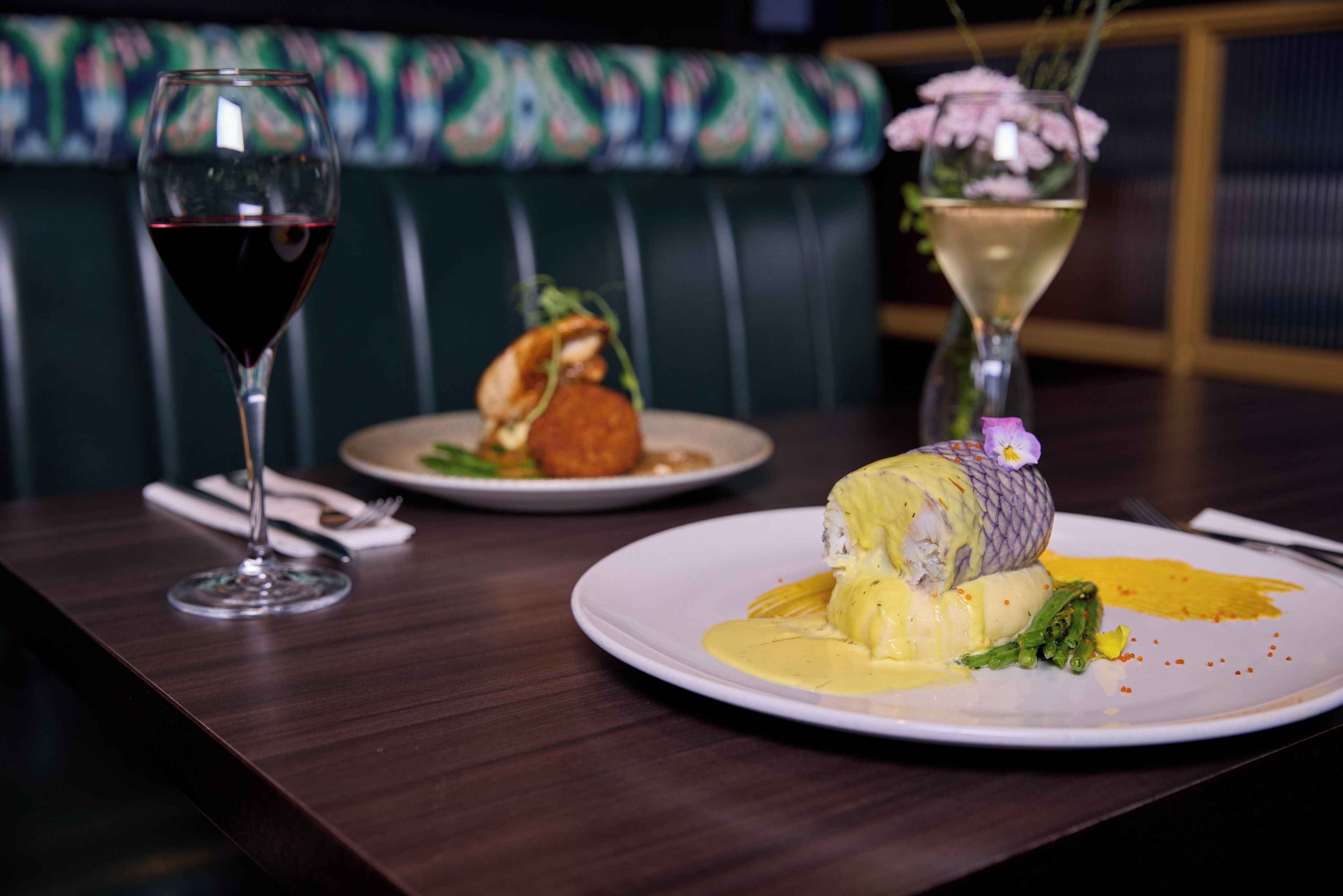 Une table à manger avec deux assiettes de nourriture, un verre à vin et un vase rempli de fleurs.